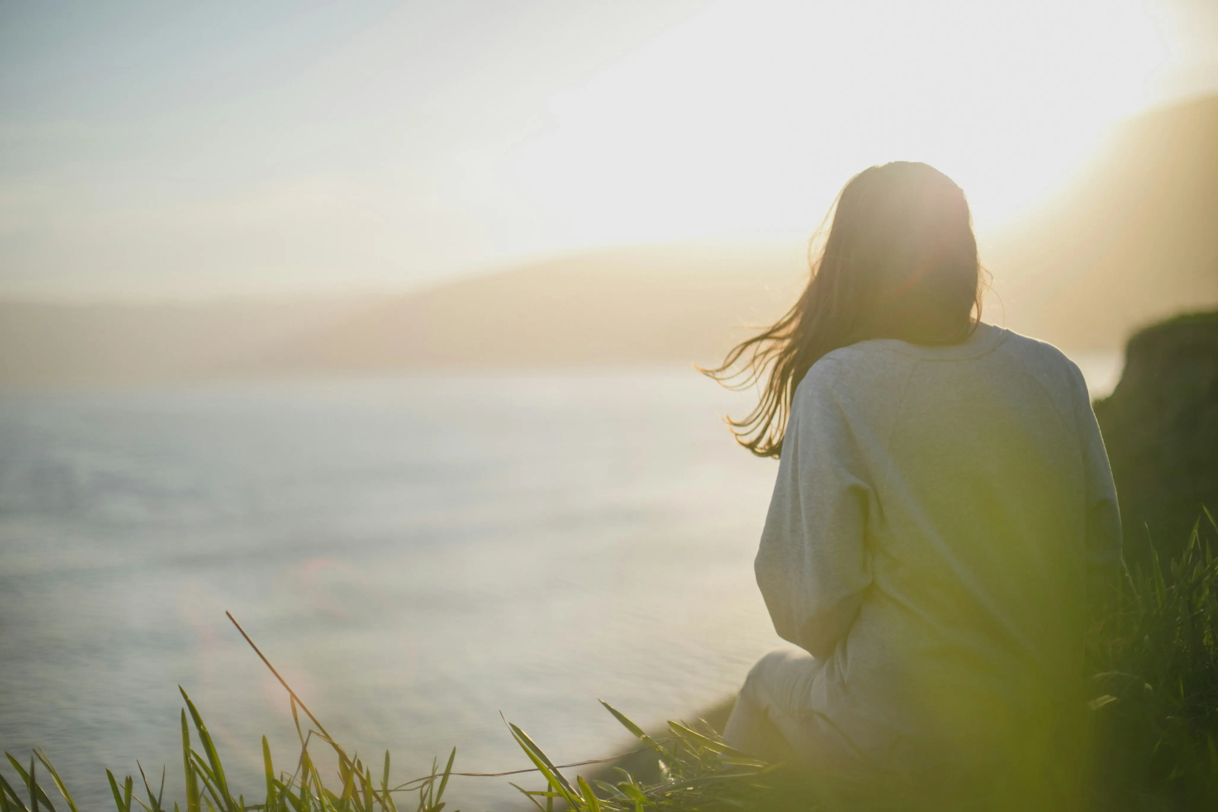 Woman sitting peacefully on a hillside overlooking a sunlit lake at dawn, representing Mind & Body Infusions' wellness blog on holistic health, longevity, and mental well-being
