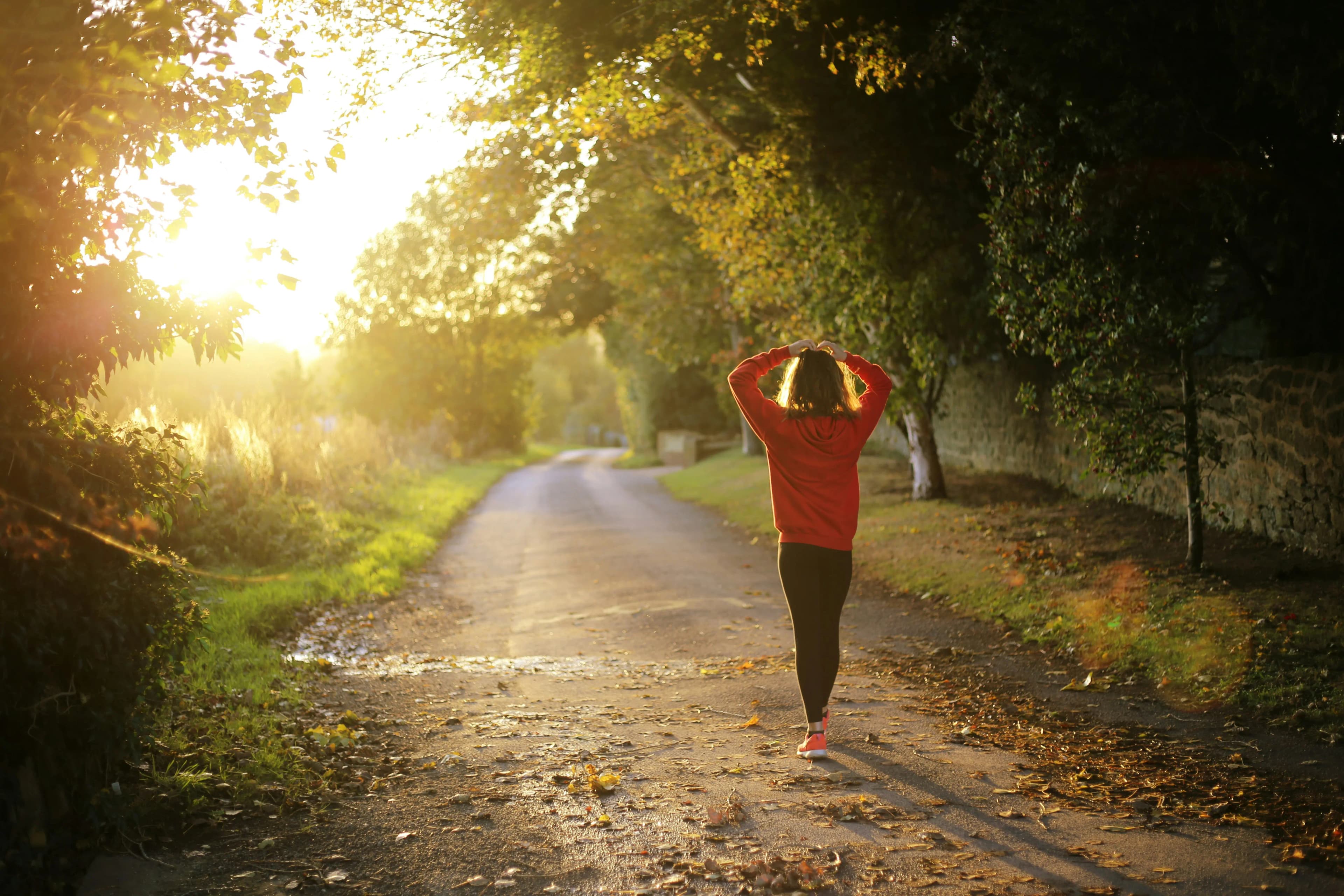 Woman in athletic wear walking on a sun-drenched wooded trail, representing the active lifestyle and vitality achieved through Mind & Body Infusions' personalized wellness care